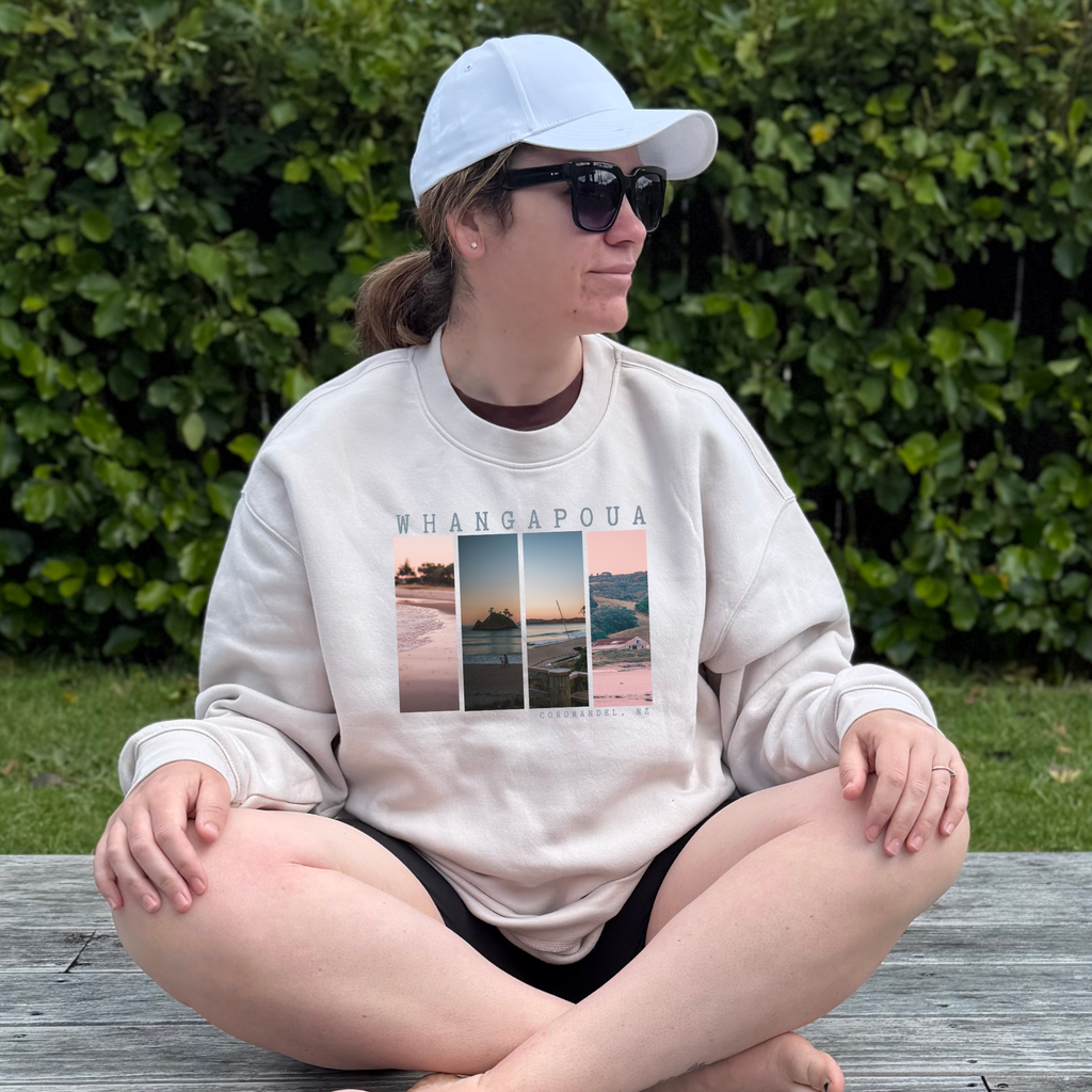 Person wearing a sweatshirt with 'Whangapoua' design sitting on a wooden bench.